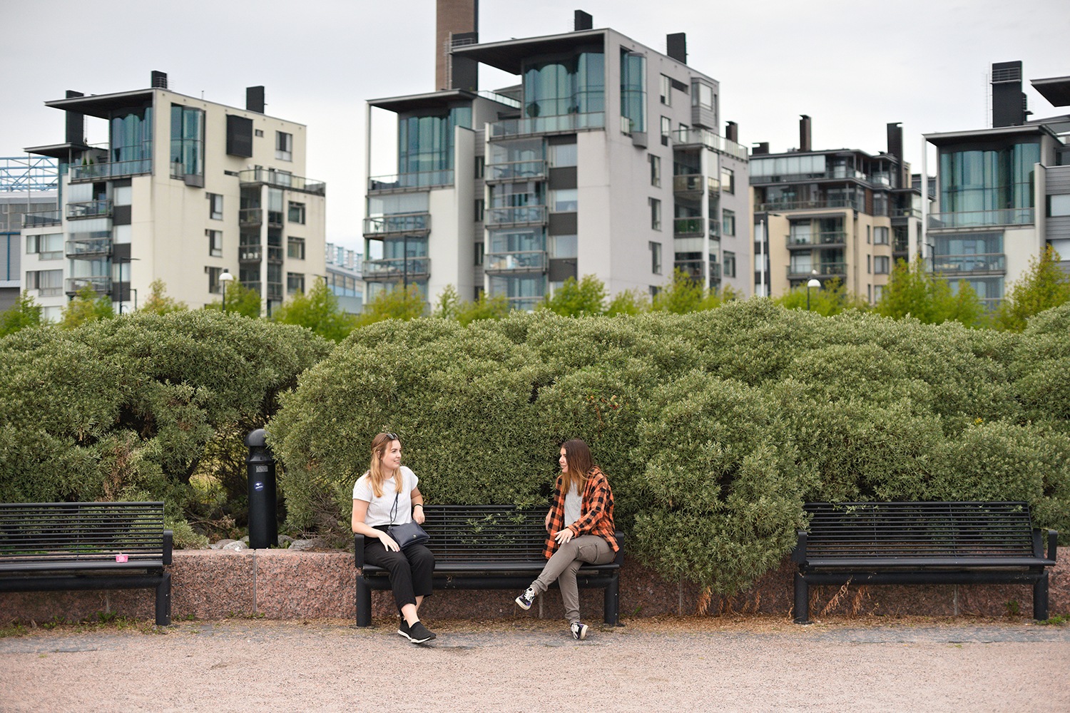 Kaksi naista istuu puistonpenkeillä Eiranrannassa vehreiden pensaiden edessä