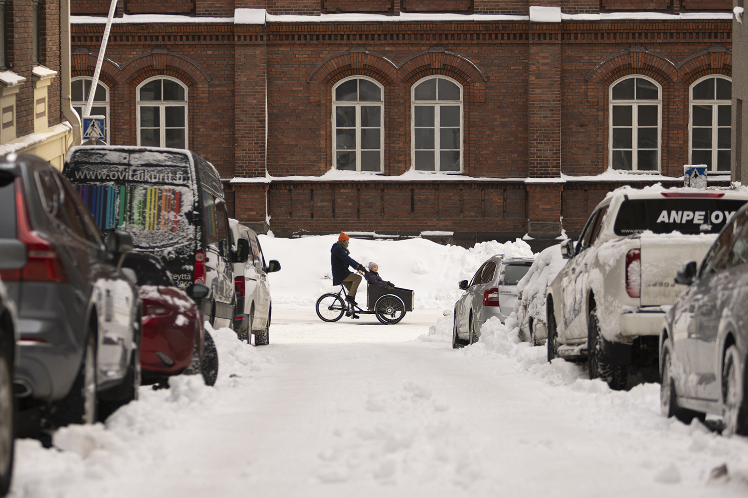 Mies ajaa lumisella kadulla kuormapyörää, jonka kyydissä on nuori tyttö.
