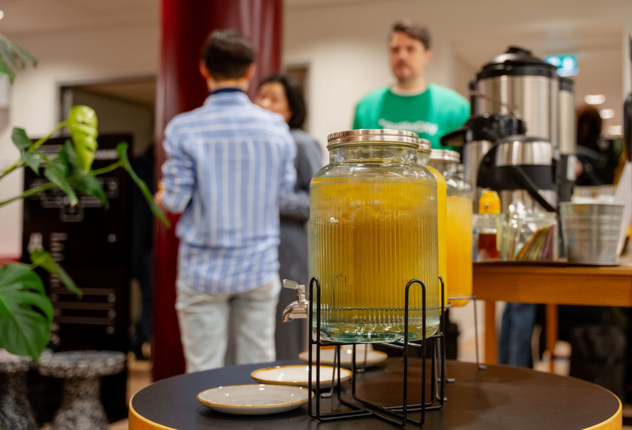 Mehuastioita täynnä keltaista mehua, iso kahviastia. Taustalla kolme henkilöä.