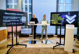 "Two men are standing in a room with tall windows in the background. On both sides of the men, there are screens on pedestals displaying text.