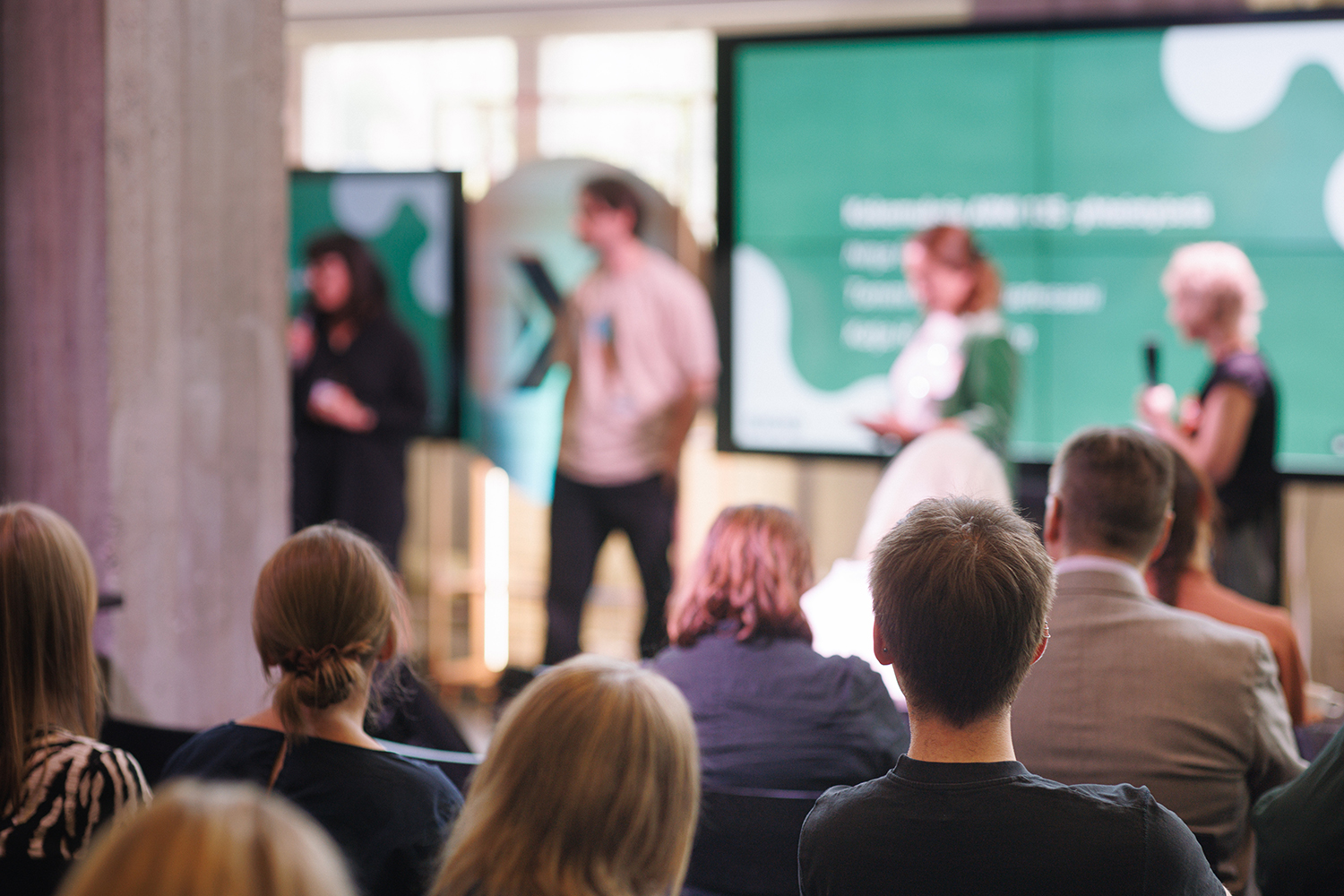 Audience members viewed from behind listening to a panel discussion, with the speakers blurred in the background.